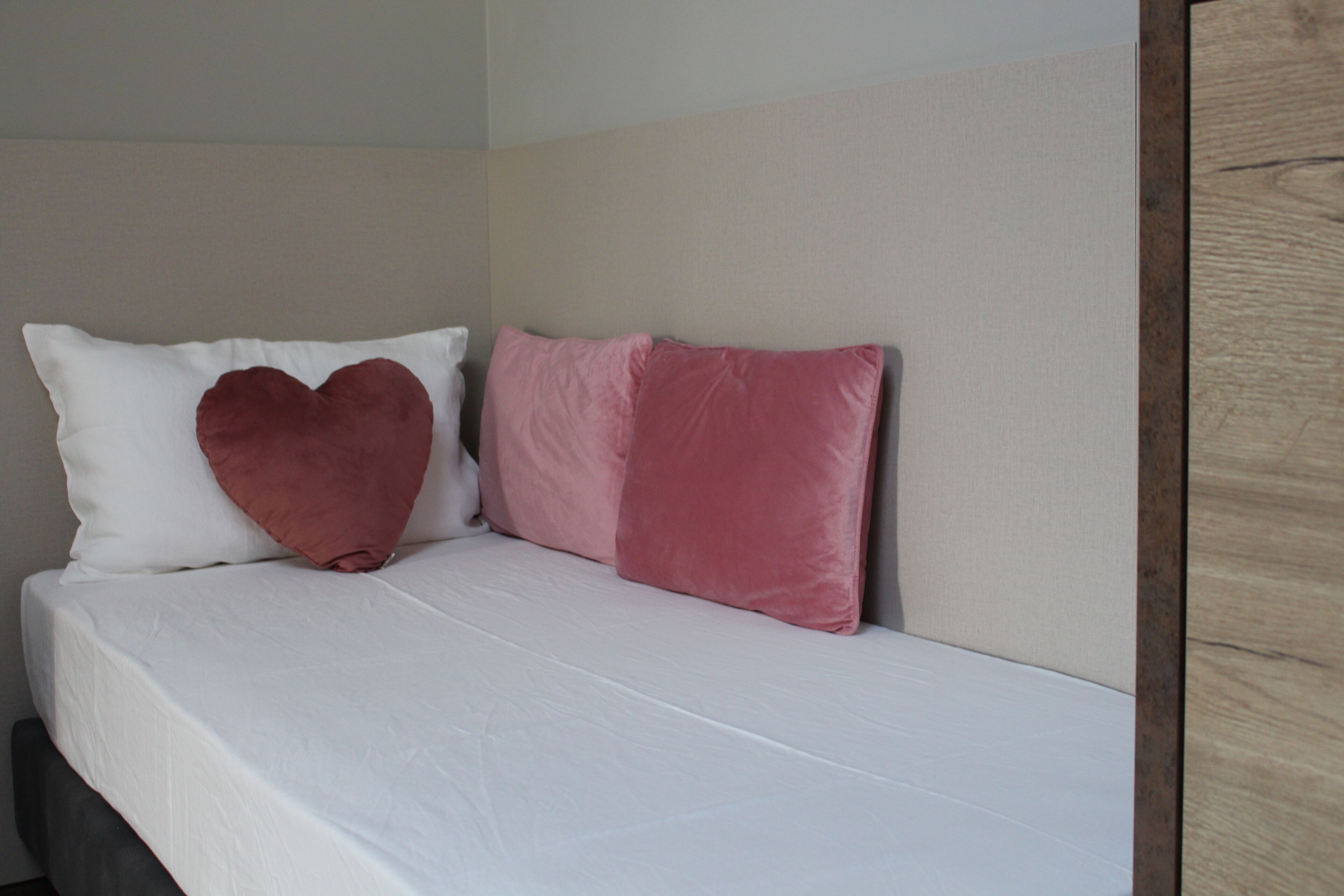 Una cama con ropa de cama blanca y dos almohadas, una con forma de corazón y otra rectangular, en una habitación con cabecera de madera y pared lisa.