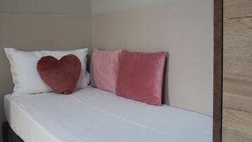 Un lit avec une literie blanche et deux oreillers, l’un en forme de cœur et l’autre rectangulaire, dans une chambre dotée d’une tête de lit en bois et d’un mur uni.