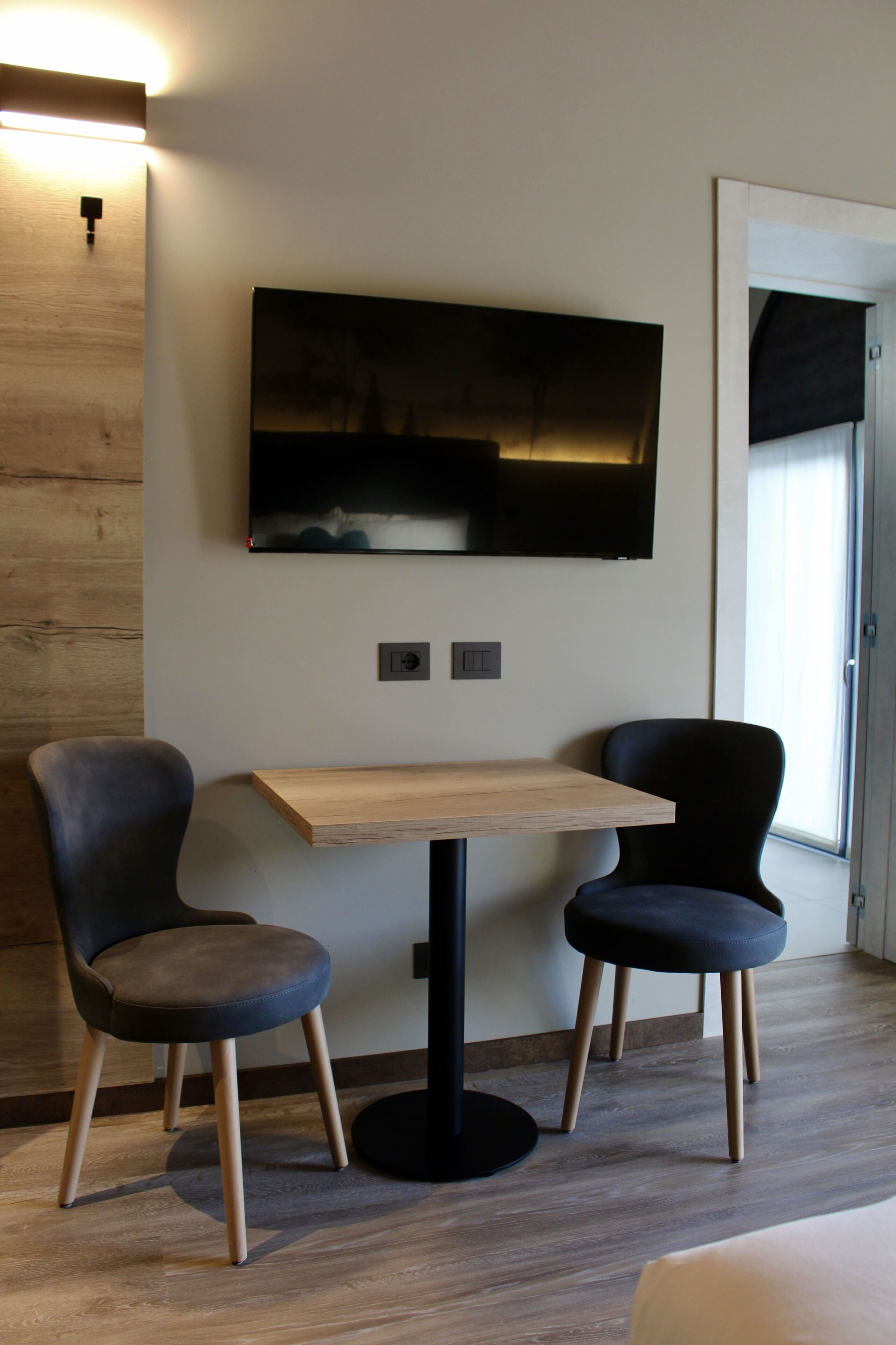 Habitación moderna con una mesa de madera, dos sillas, un televisor de pantalla plana montado en la pared y una ventana con persianas.