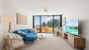 A modern living room with a blue sofa, a wicker chair, a flat-screen TV, and a balcony with a view of trees and a blue sky.