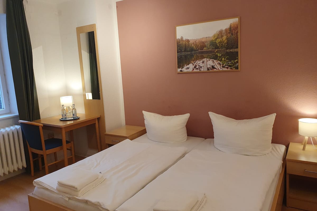 Une chambre d’hôtel avec un lit, deux oreillers, un bureau avec une chaise, un miroir, une table de chevet avec une lampe et un tableau au mur.