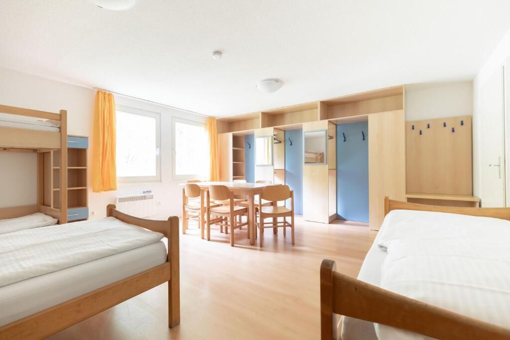 Een slaapzaal met stapelbedden, een eettafel met stoelen en een kledingkast met meerdere deuren.