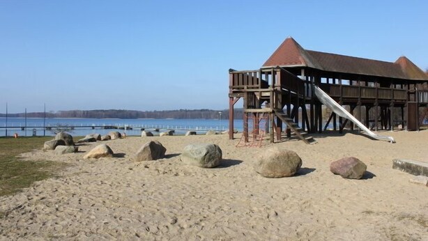 Een houten constructie met een rood dak en een trap die leidt naar een platform, gelegen op een zandstrand met grote rotsen en een waterpartij op de achtergrond.