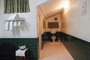 Un baño con paredes de azulejos blancos, una sección inferior de azulejos verdes, un inodoro, un lavamanos y un perchero con toallas enrolladas.
