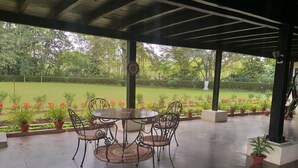 A covered patio with a round table and chairs, surrounded by potted plants and a view of a well-maintained garden.