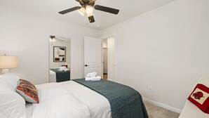 Une chambre à coucher bien rangée, avec un lit, une table de chevet, une chaise, un ventilateur de plafond et une porte donnant sur une autre pièce.