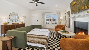 Une chambre d’hôtel moderne avec un sol à carreaux, un lit recouvert d’une courtepointe à motifs léopard, un canapé vert, un fauteuil orange, une cheminée et un ventilateur de plafond.