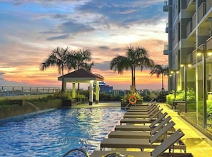 A swimming pool with lounge chairs, a gazebo, palm trees, and a sunset view.