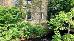 A stone building with multiple windows, surrounded by lush greenery and a small stream. - The Old Riverside School House Apt 3 (Hebden Bridge)
