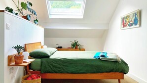 A bedroom with a skylight, a bed, a bedside table, a chair, a small table, a potted plant, and a painting on the wall. - The Old Riverside School House Apt 3 (Hebden Bridge)