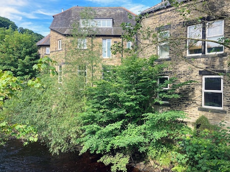 A stone building with multiple windows, surrounded by dense greenery and a small stream.