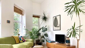 A living room with a green sofa, a flat-screen TV, a coffee table, and several potted plants. - The Old Riverside School House Apt 3 (Hebden Bridge)