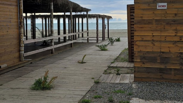 A wooden walkway leading to a beach with thatched-roof structures and a wooden wall labeled "SHOWER". - Tirent Apartment - Cozy Bedrooms Stay by Golem Beach. (Golem)