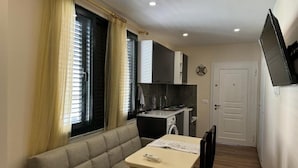 A compact living space with a sofa, a dining table, a kitchen area with a washing machine, and a TV mounted on the wall. - Tirent Apartment - Cozy Bedrooms Stay by Golem Beach. (Golem)