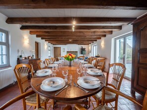 Une salle à manger avec une longue table en bois dressée pour un repas, entourée de chaises en bois, et un vase rempli de fleurs placé au centre.