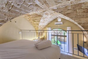 A bedroom with a high, arched stone ceiling, a bed with white linens, a balcony with a railing, and a view of greenery outside. - Casa Batroun Lofts (Batroun)