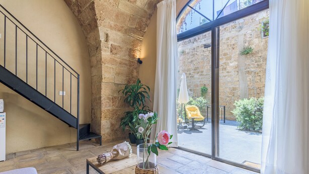 Ein geräumiges Zimmer mit Steinwand, einem Holztisch mit Blumenvase, einer Treppe und Blick auf den Innenhof durch große Glastüren.