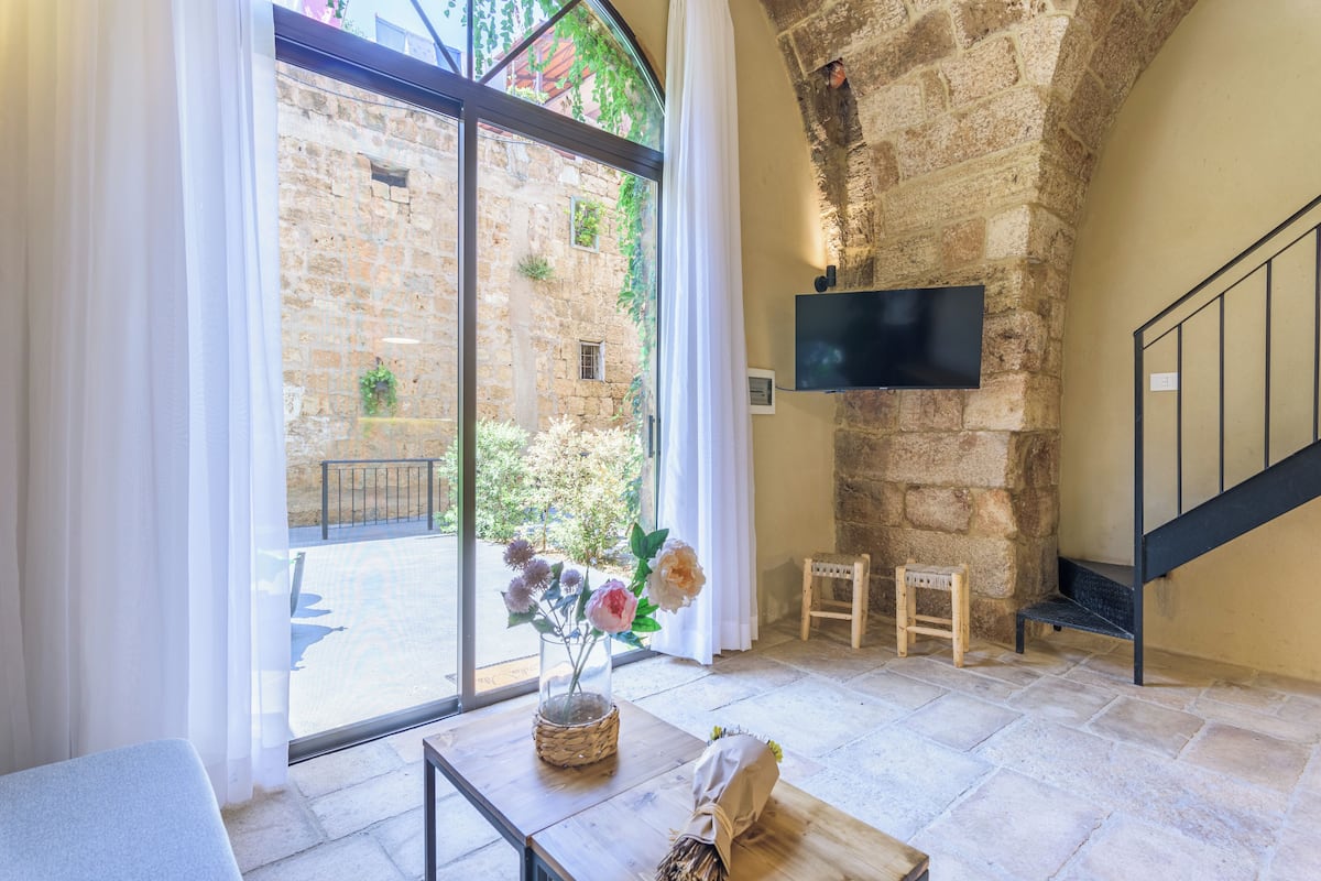 Une pièce avec un mur en pierre, un téléviseur à écran plat fixé au mur, un banc en bois et une petite table sur laquelle est posé un vase de fleurs.