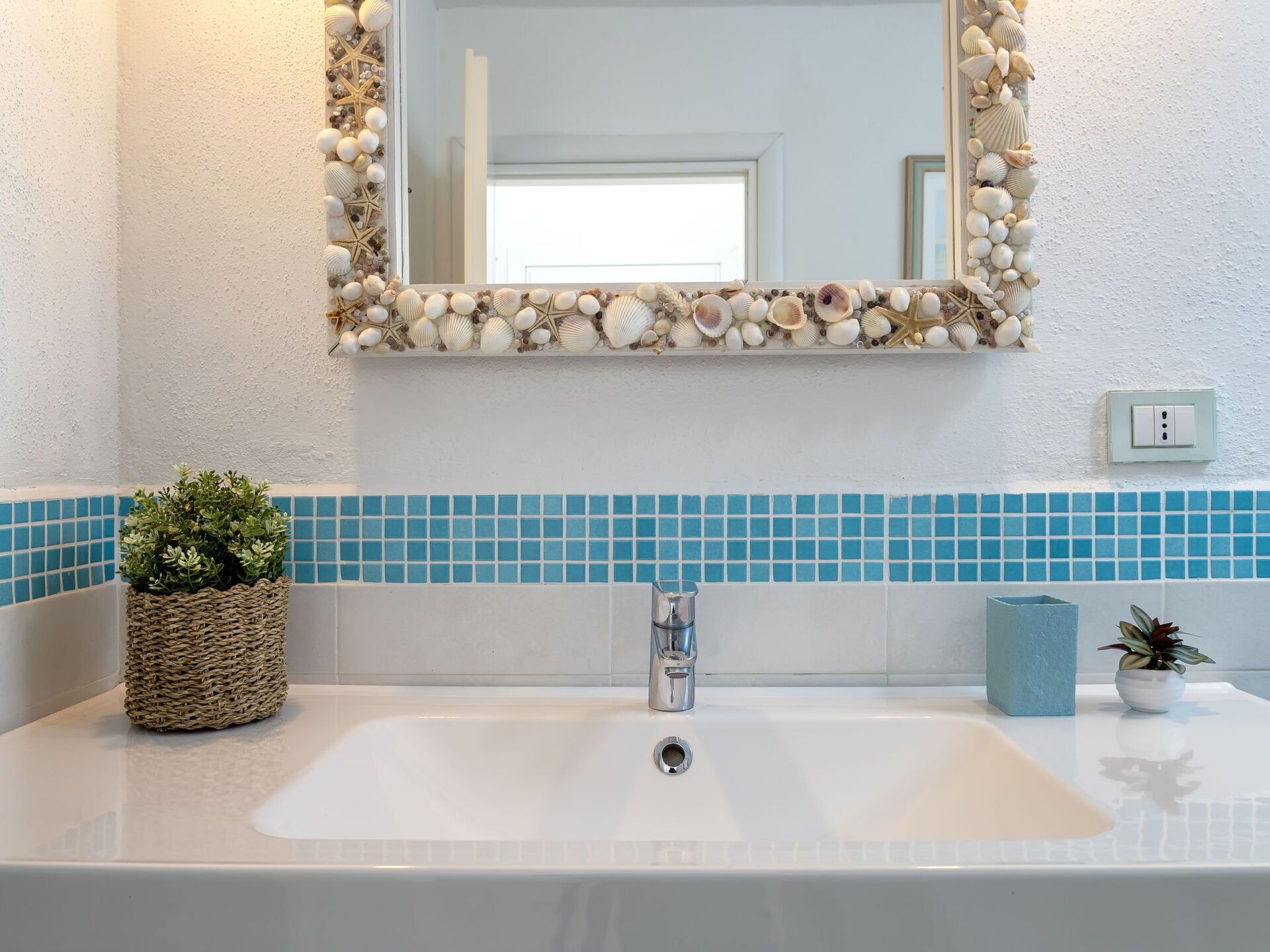 Ein Badezimmer mit einem weißen Waschbecken, blauen Mosaikfliesen, einem Spiegel mit einer Muschelgirlande und einer kleinen Pflanze.