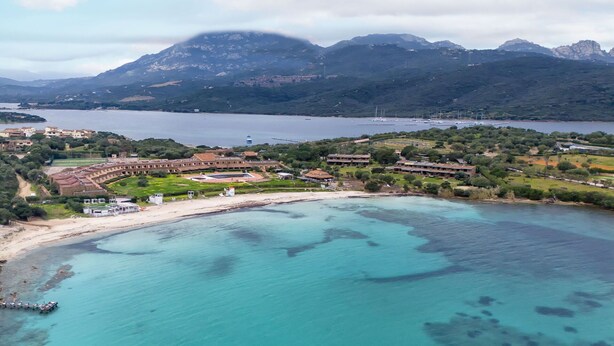 Vista aèria d'un complex turístic costaner amb platja, viles i un paisatge de muntanyes al fons.