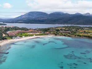 Vista aérea de un complejo turístico costero con playa, villas y montañas al fondo.