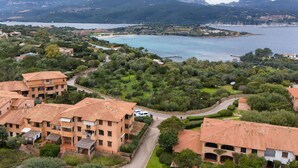 Zona residencial a la costa amb edificis de teulades de terracota, vegetació exuberant i una vista al mar i a les muntanyes de fons.