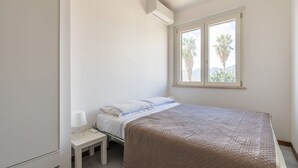 Un dormitorio con cama, mesita de noche, lámpara y ventana con vistas a palmeras.