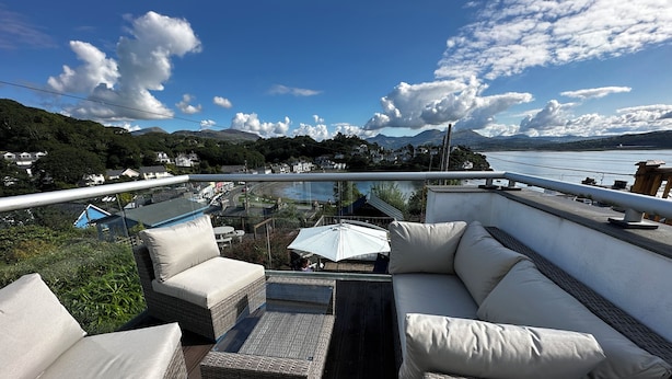 A balcony with wicker furniture and a glass railing overlooking a scenic landscape with mountains and a body of water. - Alltwen (Porthmadog)