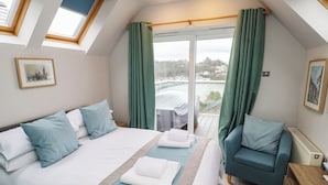 A bedroom with a large bed, a blue armchair, and a view of a body of water through a sliding glass door. - Alltwen (Porthmadog)