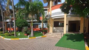A paved courtyard with a green lawn, palm trees, and a building entrance. - Millsview hotel (Kisumu)