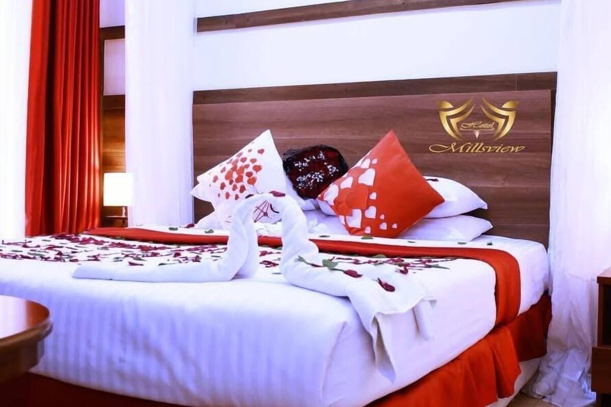 A hotel room with a neatly made bed, red curtains, and a wooden headboard with the hotel logo.