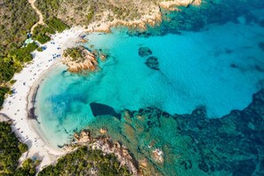 Vue aérienne d’une plage aux eaux turquoise et limpides, avec des affleurements rocheux et une côte sablonneuse.