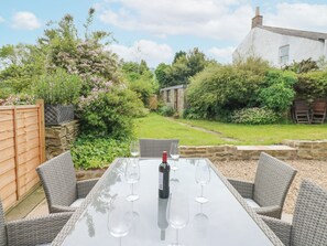 An outdoor dining area with wicker chairs and a glass table, a bottle of wine, and a view of a garden with a stone wall and a white building in the background. - Valley View (Leyburn)