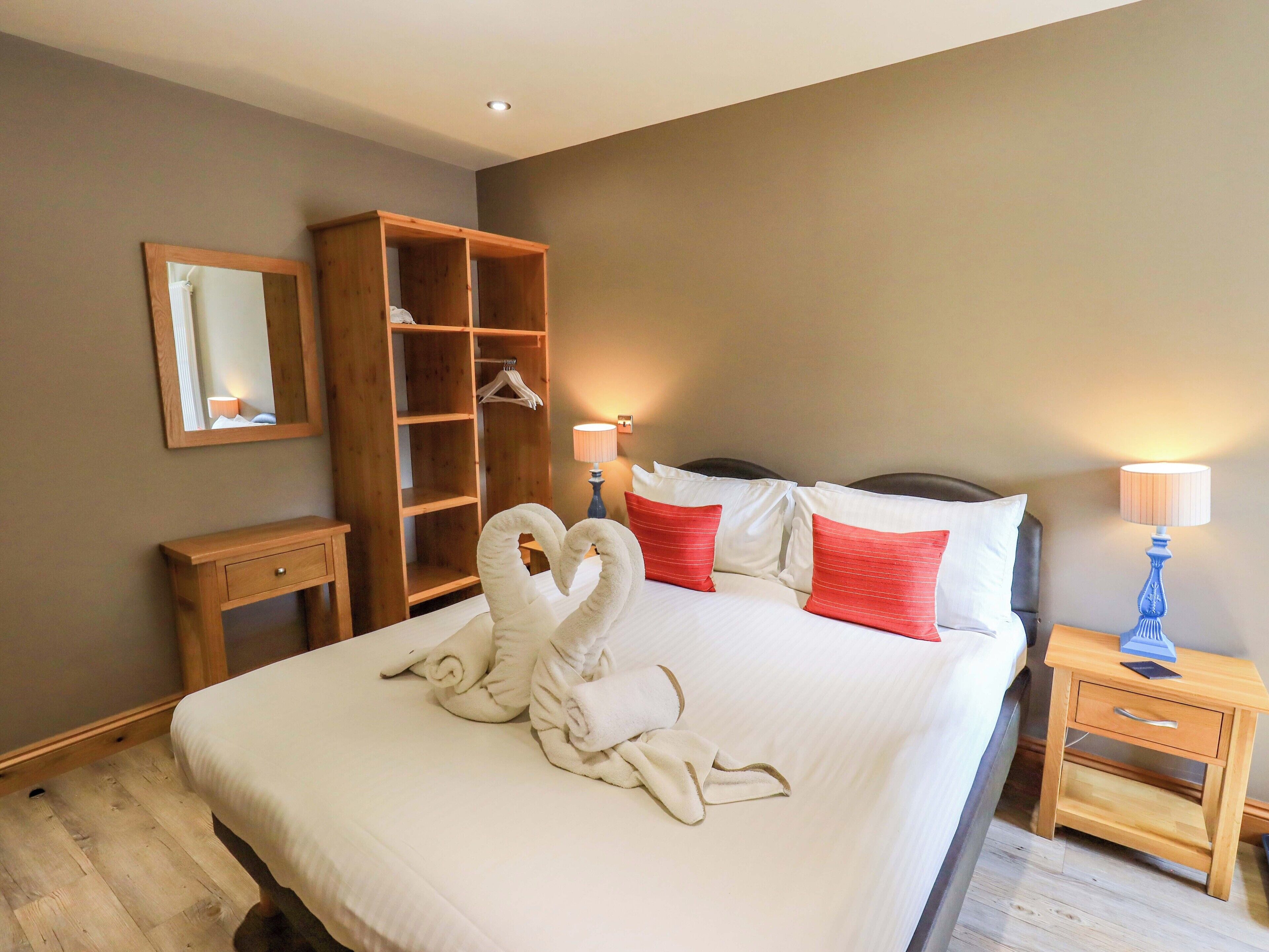 A hotel room with a large bed, a wooden wardrobe, a bedside table with a lamp, and a mirror on the wall.