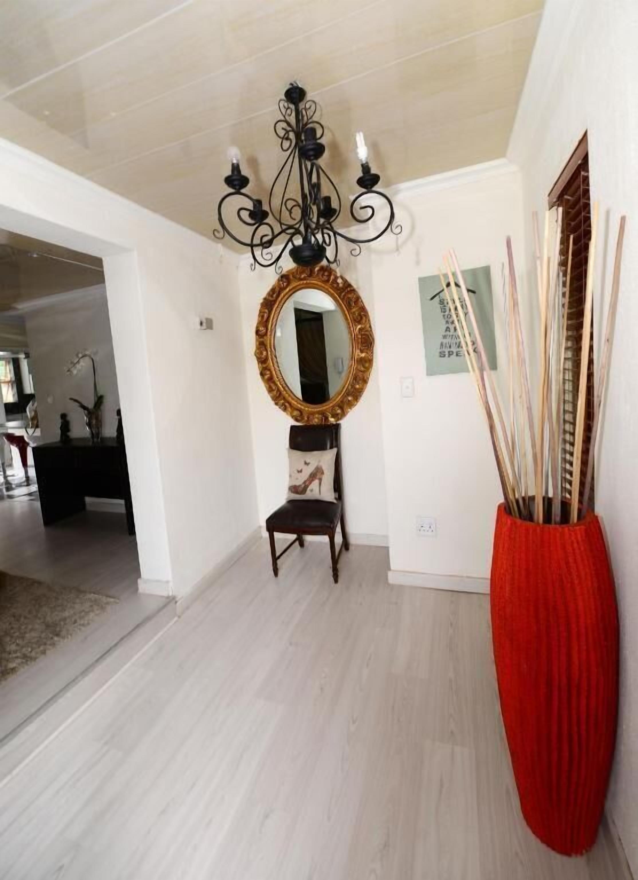 A hallway with a chandelier, a mirror, a chair, and a red vase with sticks.