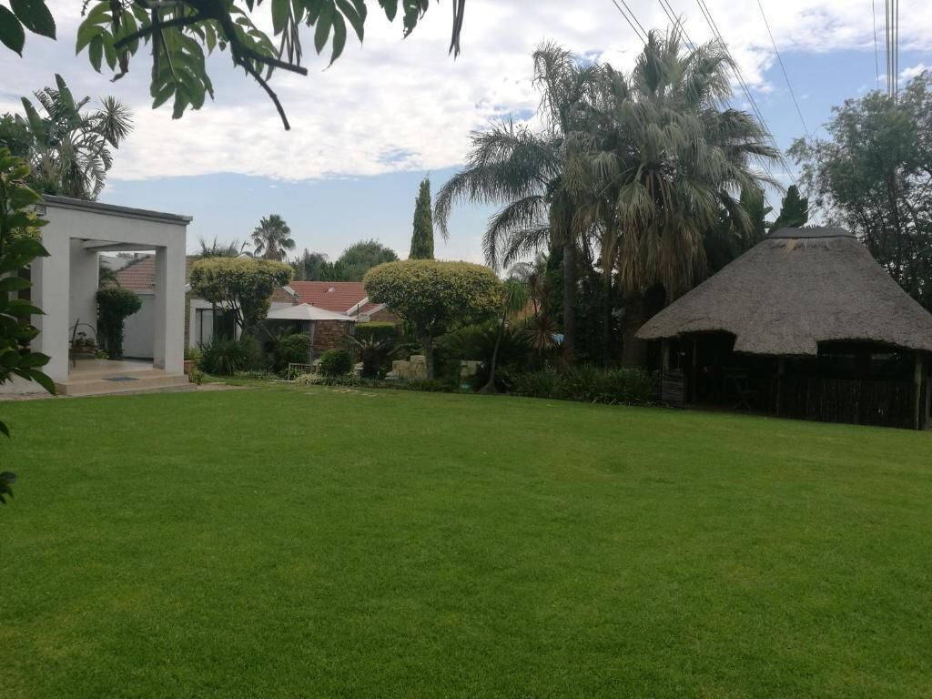 A well-maintained lawn with a thatched-roof structure, a white building with an archway, and various trees and shrubs.