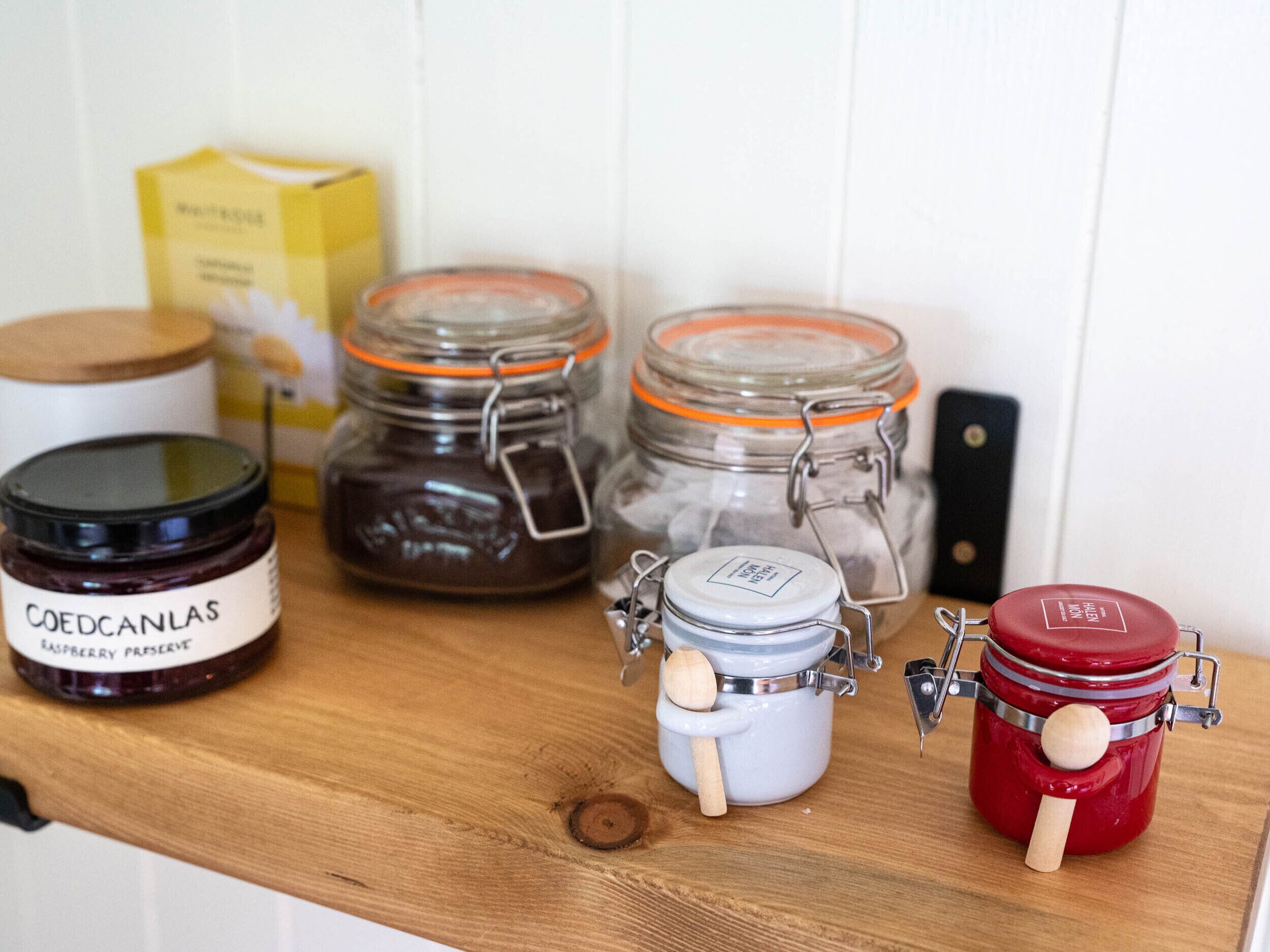 A wooden shelf with various jars of preserves, including a raspberry preserve and a honey preserve.
