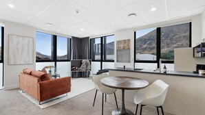 Een moderne hotelkamer met een groot raam met uitzicht op de bergen, een gezellige zithoek met een bruine bank, een ronde eettafel met twee stoelen, en een keuken met een aanrecht en spoelbak.