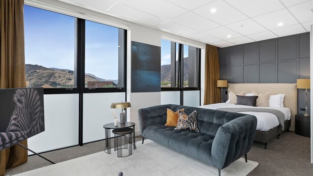Une chambre d’hôtel moderne avec un grand lit, un canapé et un bureau. De grandes fenêtres offrent une vue sur l’extérieur.