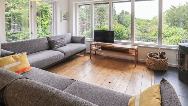 A modern living room with a grey sectional sofa, a wooden coffee table, a flat-screen TV, and large windows offering a view of greenery. - Tower Cottage (Windermere)