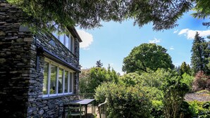 A stone building with a patio area featuring a table and chairs, surrounded by lush greenery and trees. - Tower Cottage (Windermere)