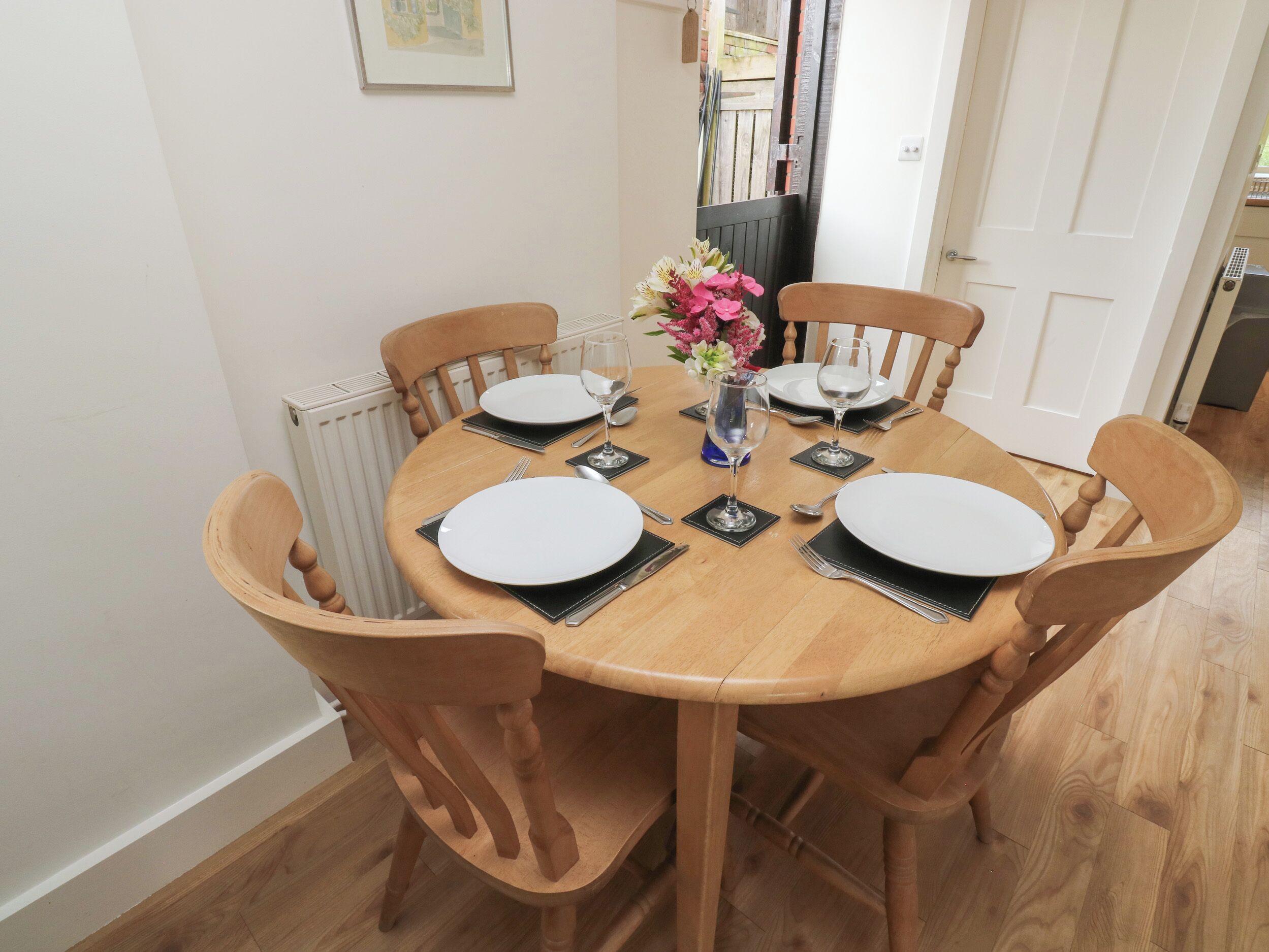 A round wooden dining table with four chairs, white plates, black napkins, and a vase with flowers.