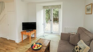 A living room with a television, a wooden coffee table with a bowl of fruit, a brown sofa, and a sliding glass door leading to a garden. - Bantam Cottage (Honiton)