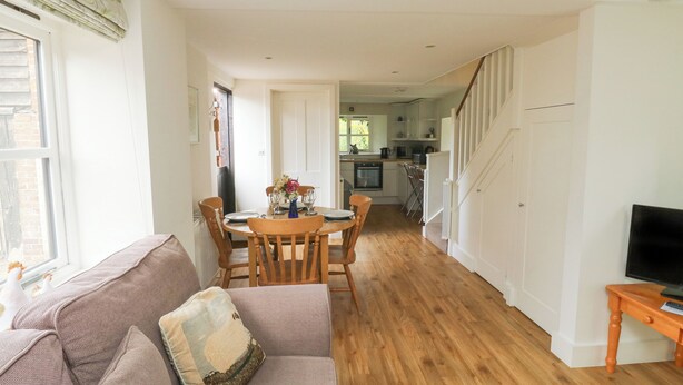A spacious living room with a sofa, a dining table set for four, and a kitchen in the background. - Bantam Cottage (Honiton)