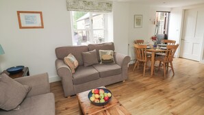 A cozy living room with a sofa, a wooden coffee table with a bowl of fruit, and a dining area with a table and chairs. - Bantam Cottage (Honiton)