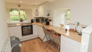 A modern kitchen with white cabinets, a wooden countertop, a built-in oven, a microwave, and a toaster. There are two bar stools and a window with a view of greenery outside. - Bantam Cottage (Honiton)