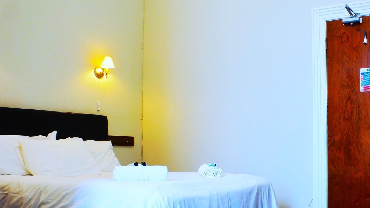 A hotel room with a bed, a nightstand, a lamp, and a wooden door.