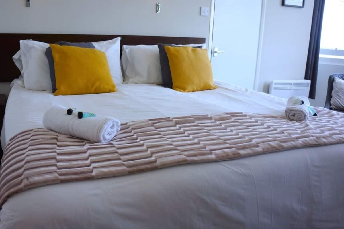 Un lit bien fait, avec une couverture à motifs, des oreillers blancs et jaunes, et une serviette enroulée posée sur le côté.