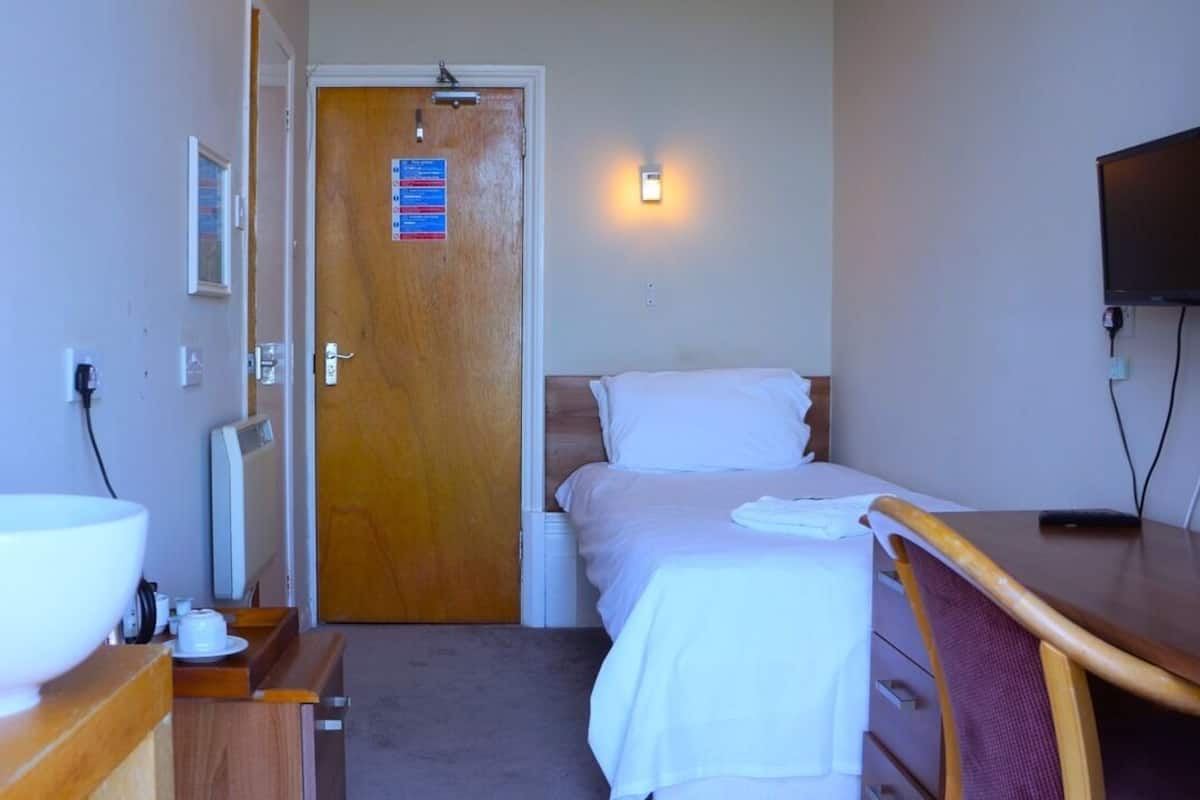 Une petite chambre d’hôtel avec un lit simple, un bureau, une chaise, une télévision et une salle de bain équipée d’un lavabo et d’un miroir.