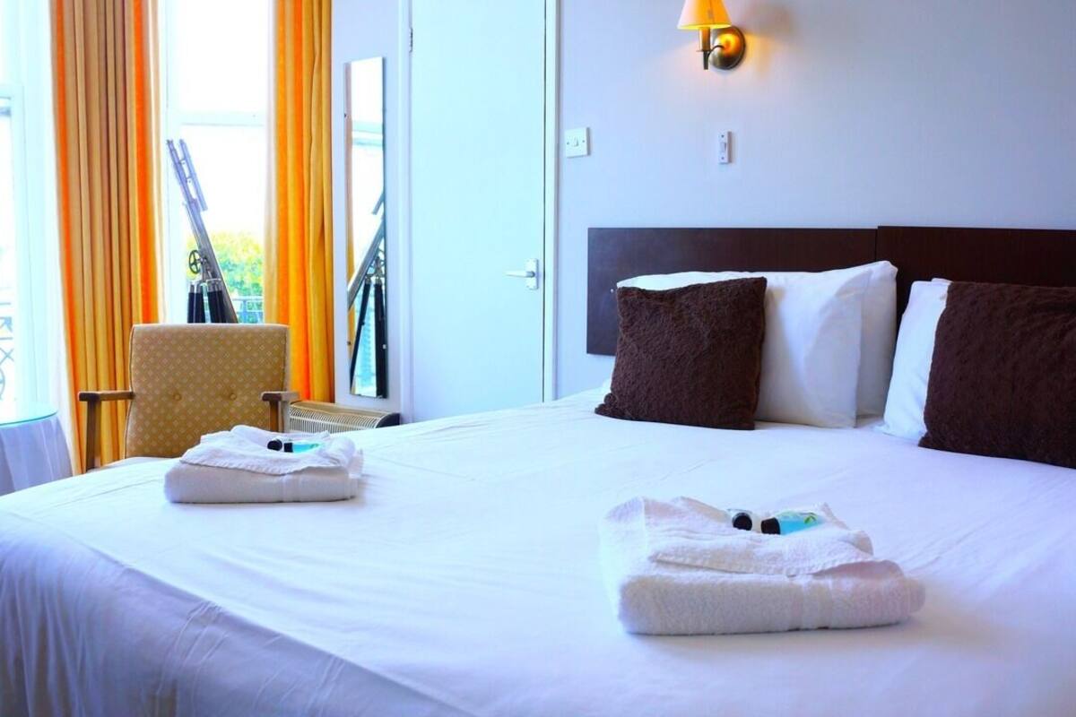 Une chambre d’hôtel avec un grand lit, deux oreillers, une chaise, une table et une fenêtre avec des rideaux.